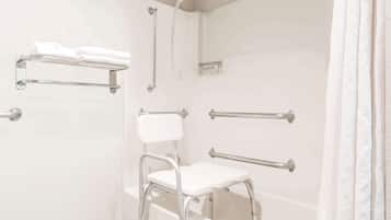 A bathroom with a shower, grab bars, a white chair, and a towel rack.