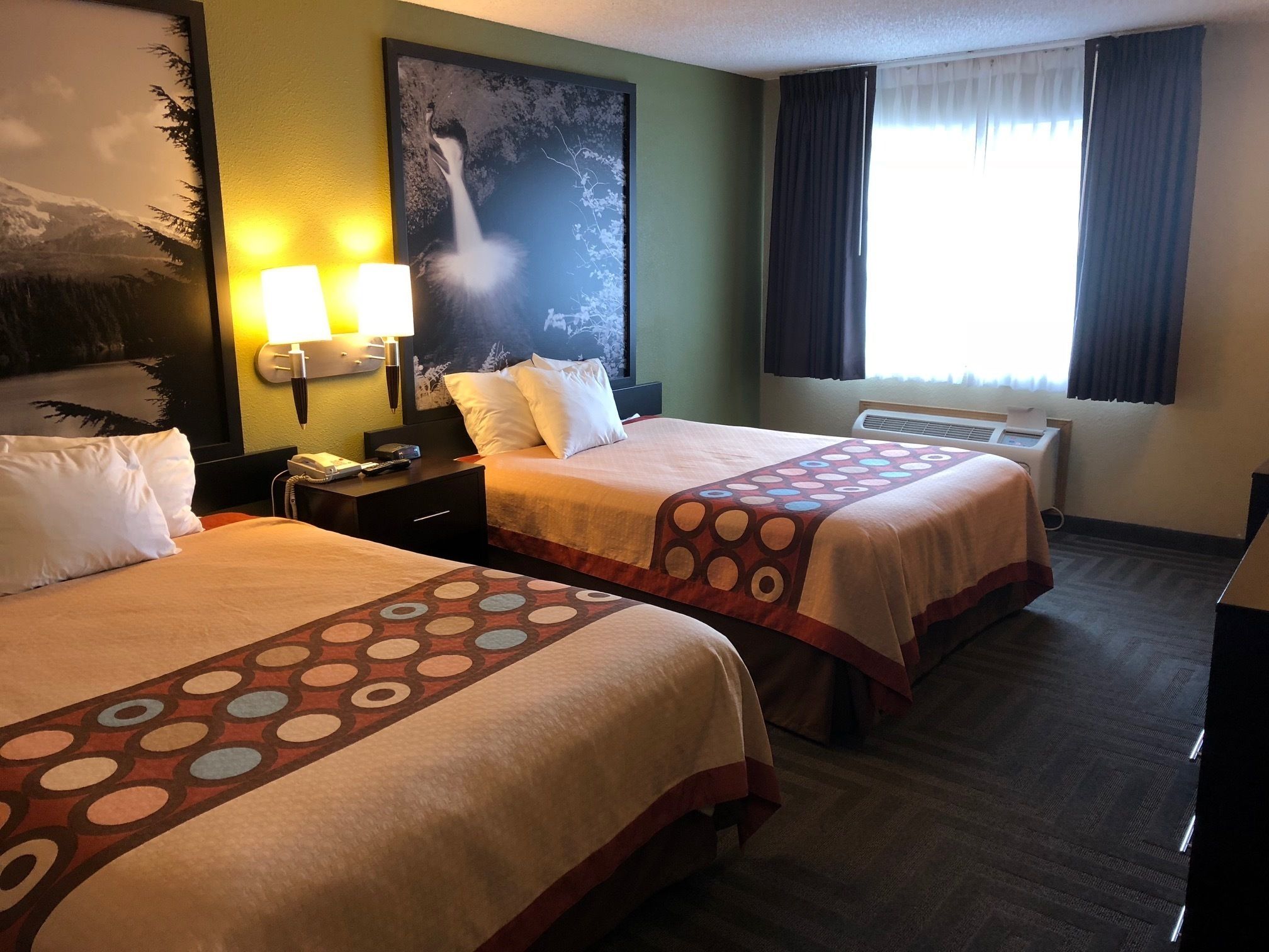 A hotel room with two beds, a nightstand, a lamp, a painting, and a window with curtains.
