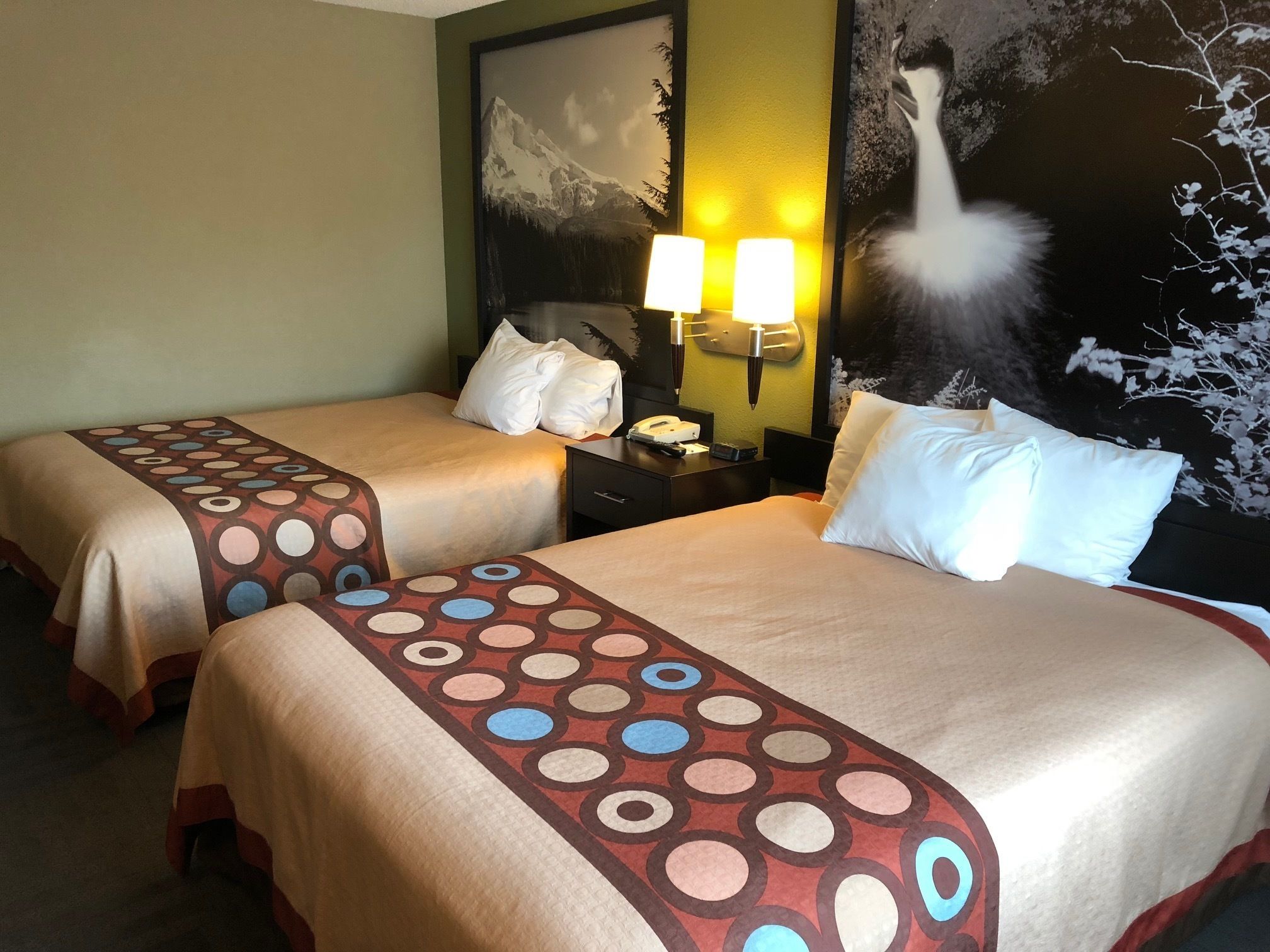 Una habitación de hotel con dos camas, cada una con una colcha estampada, una mesita de noche con lámpara y una pared decorada con un cuadro grande de temática natural.