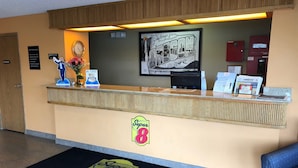 A hotel reception area with a reception desk, a clock, a framed welcome sign, and a Super 8 logo on the floor mat. - Super 8 by Wyndham LaGrande (La Grande)