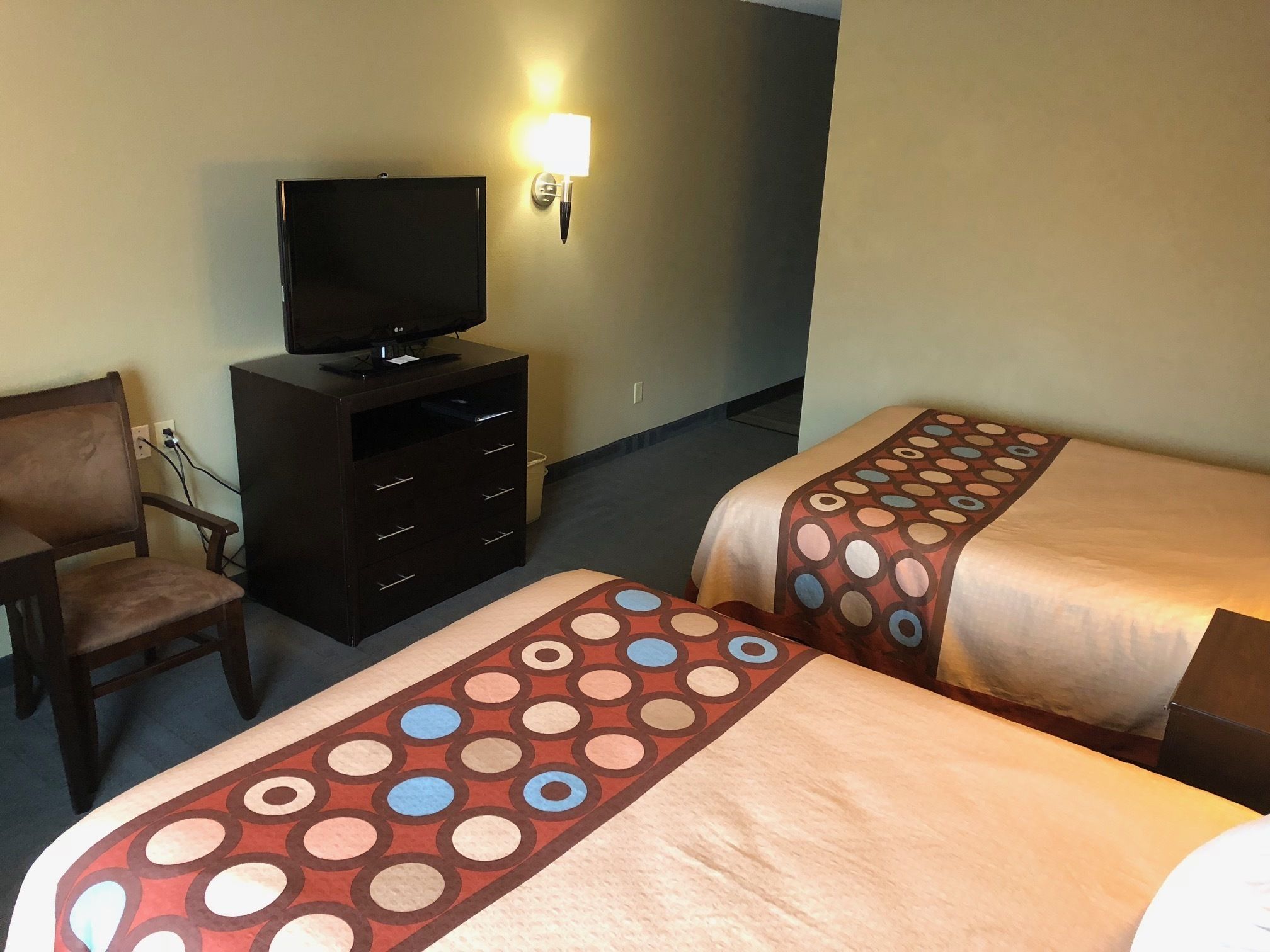 A hotel room with two beds, a TV on a dresser, a chair, and a wall-mounted lamp.