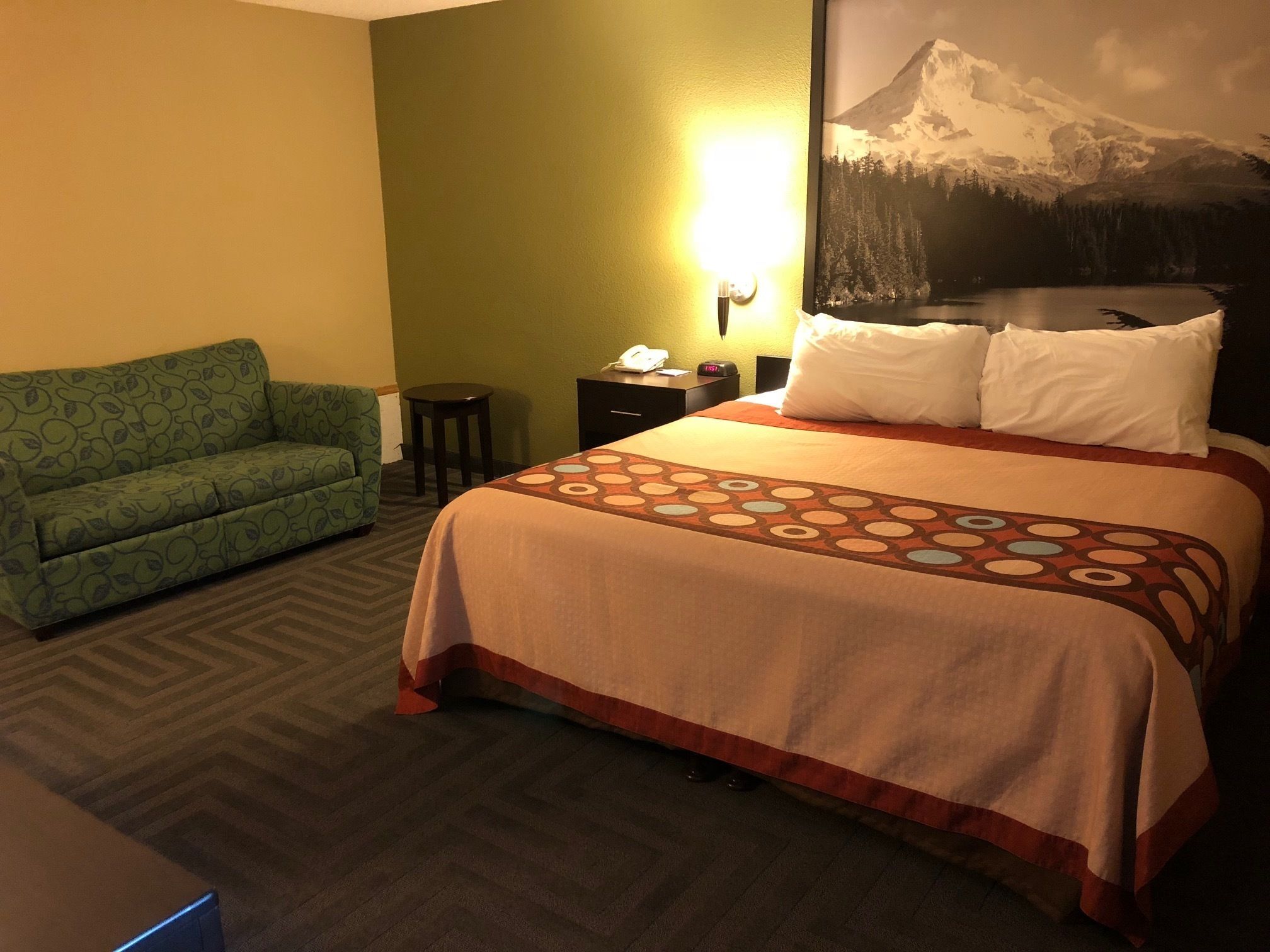 Una habitación de hotel con cama, sofá verde, mesita de noche y lámpara de pared.