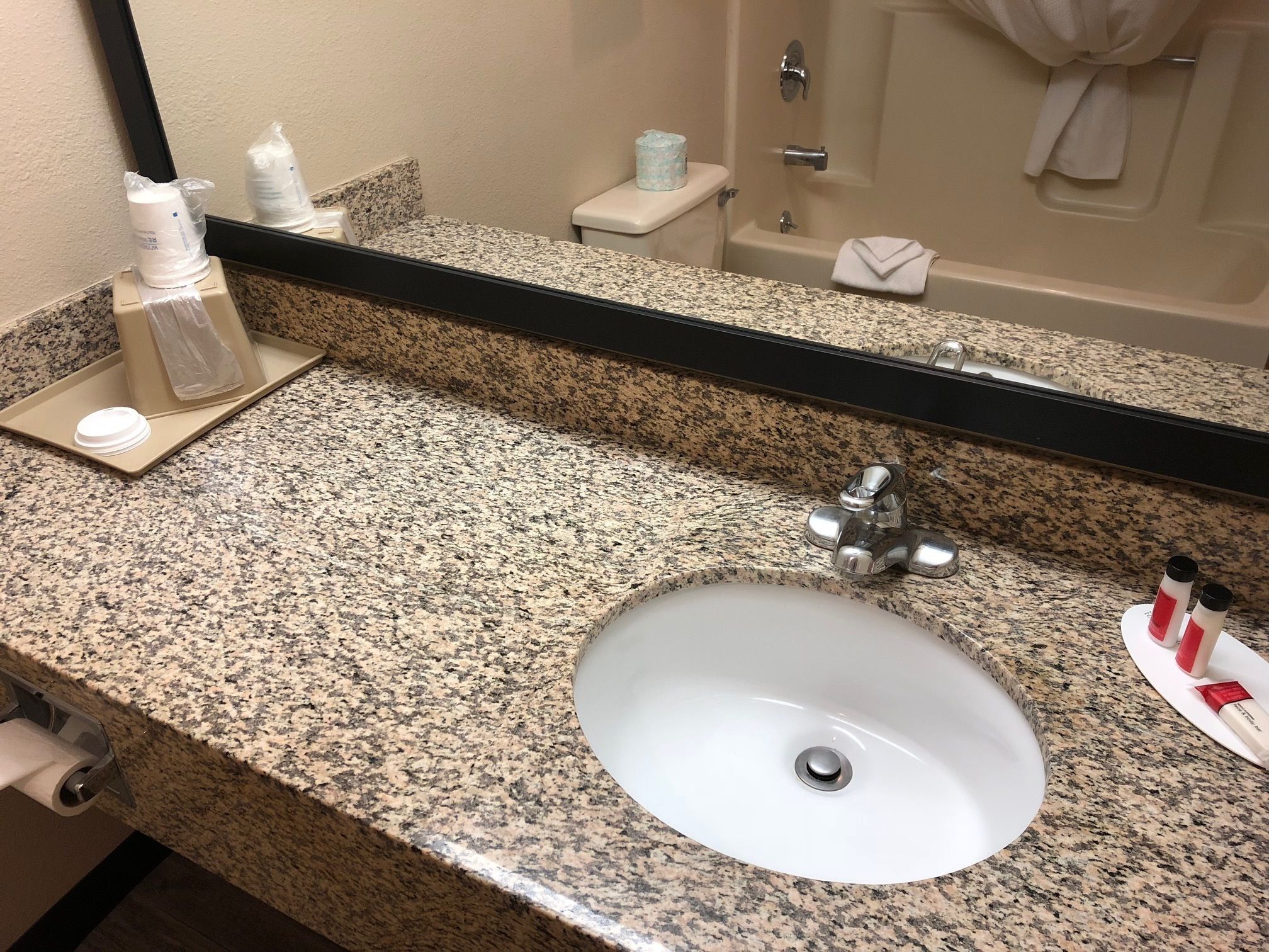 Un baño con encimera de granito, un lavabo blanco, un espejo y artículos de aseo.