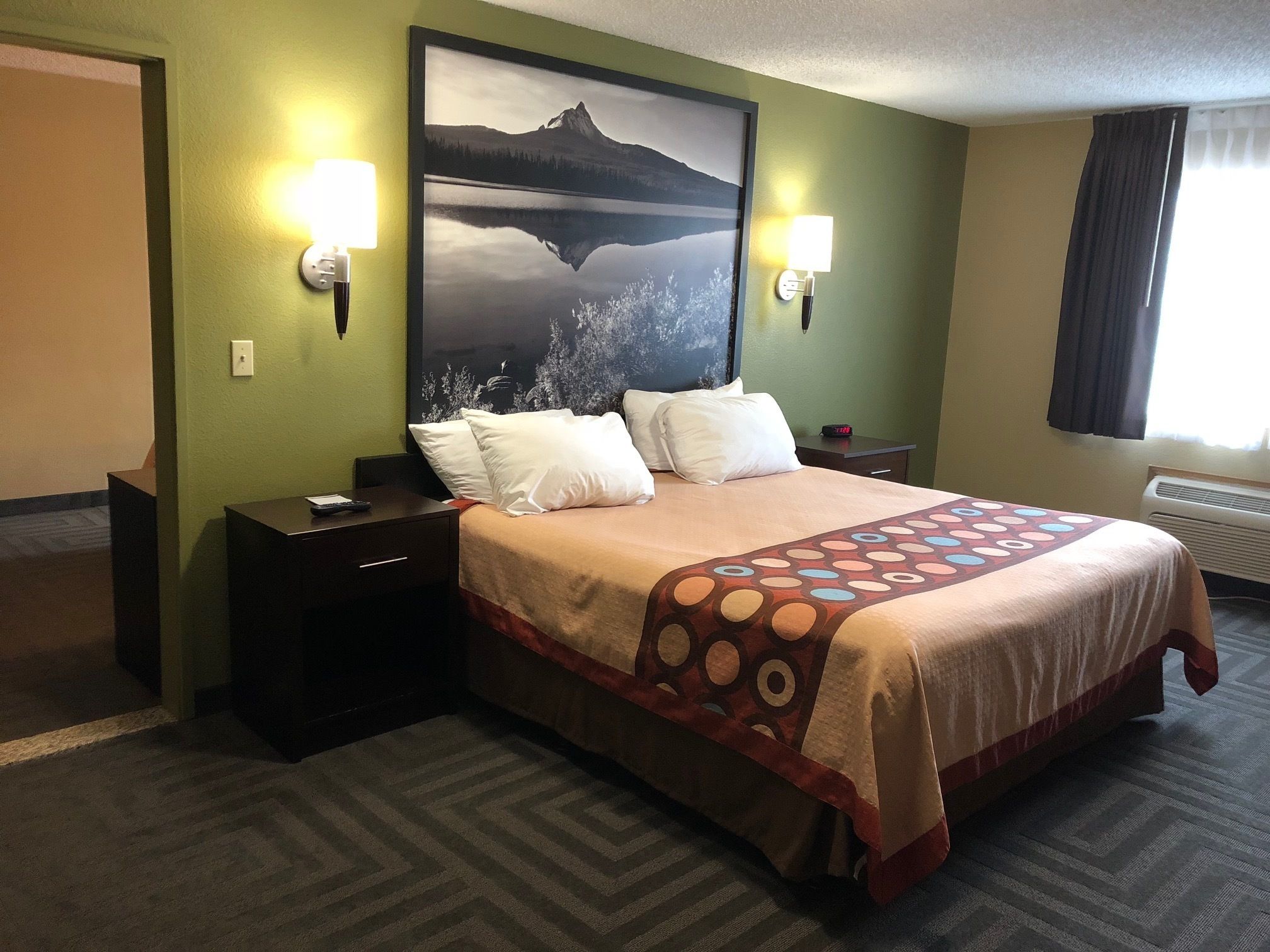Habitación de hotel con una cama, dos mesitas de noche, una lámpara de pared, un reloj y una ventana con cortinas.