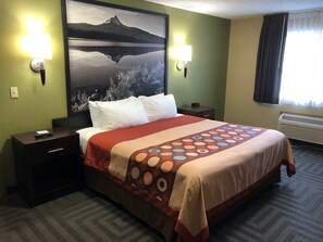 A hotel room with a bed, two bedside tables, a nightstand, a wall-mounted clock, and a large framed picture of a mountain landscape. - Super 8 by Wyndham LaGrande (La Grande)