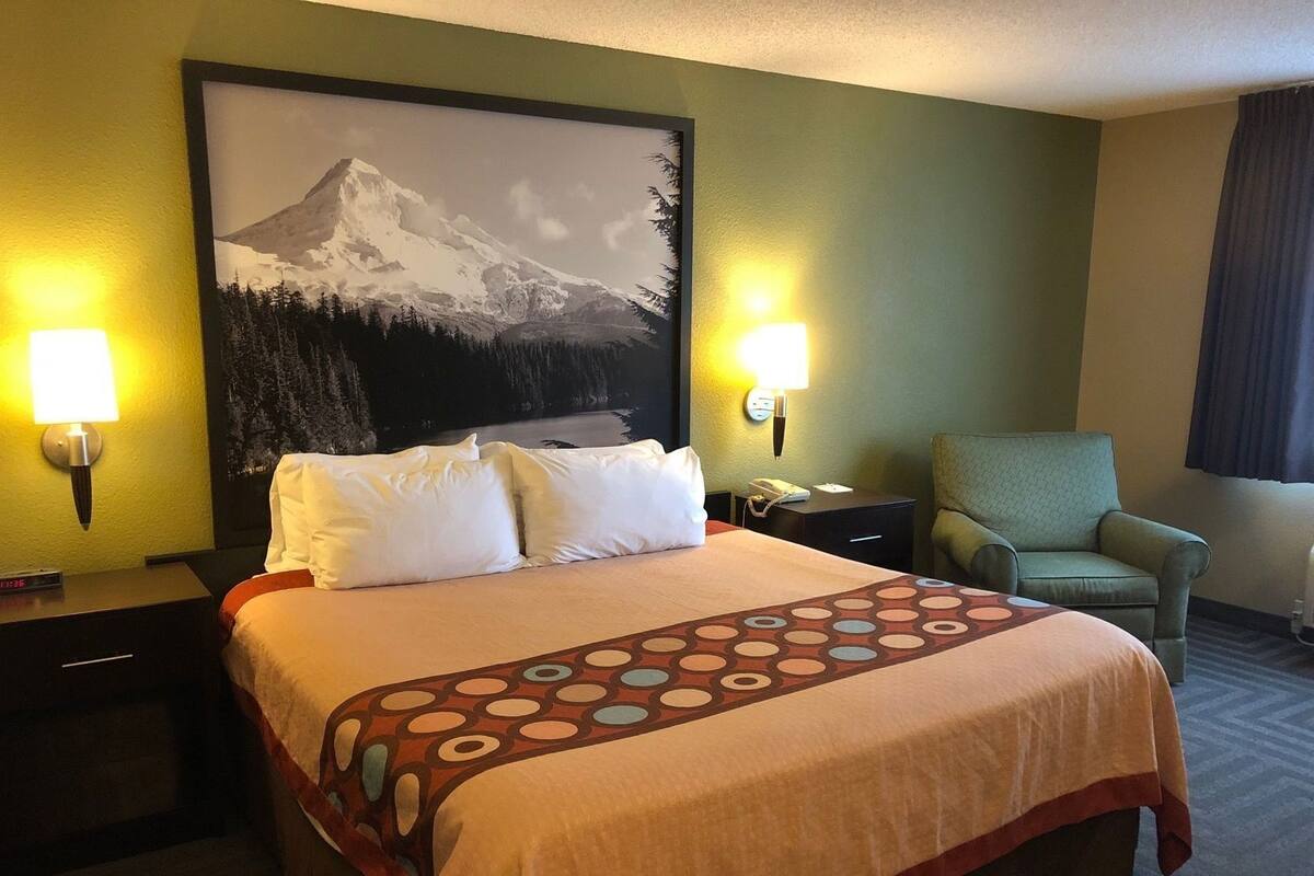 A hotel room with a bed, two bedside lamps, a chair, a painting of a mountain, and a telephone on the nightstand.