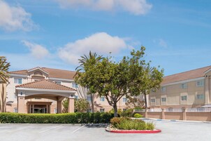 Un hotel con vestíbulo cubierto, aparcamiento y zona ajardinada con un árbol y arbustos.