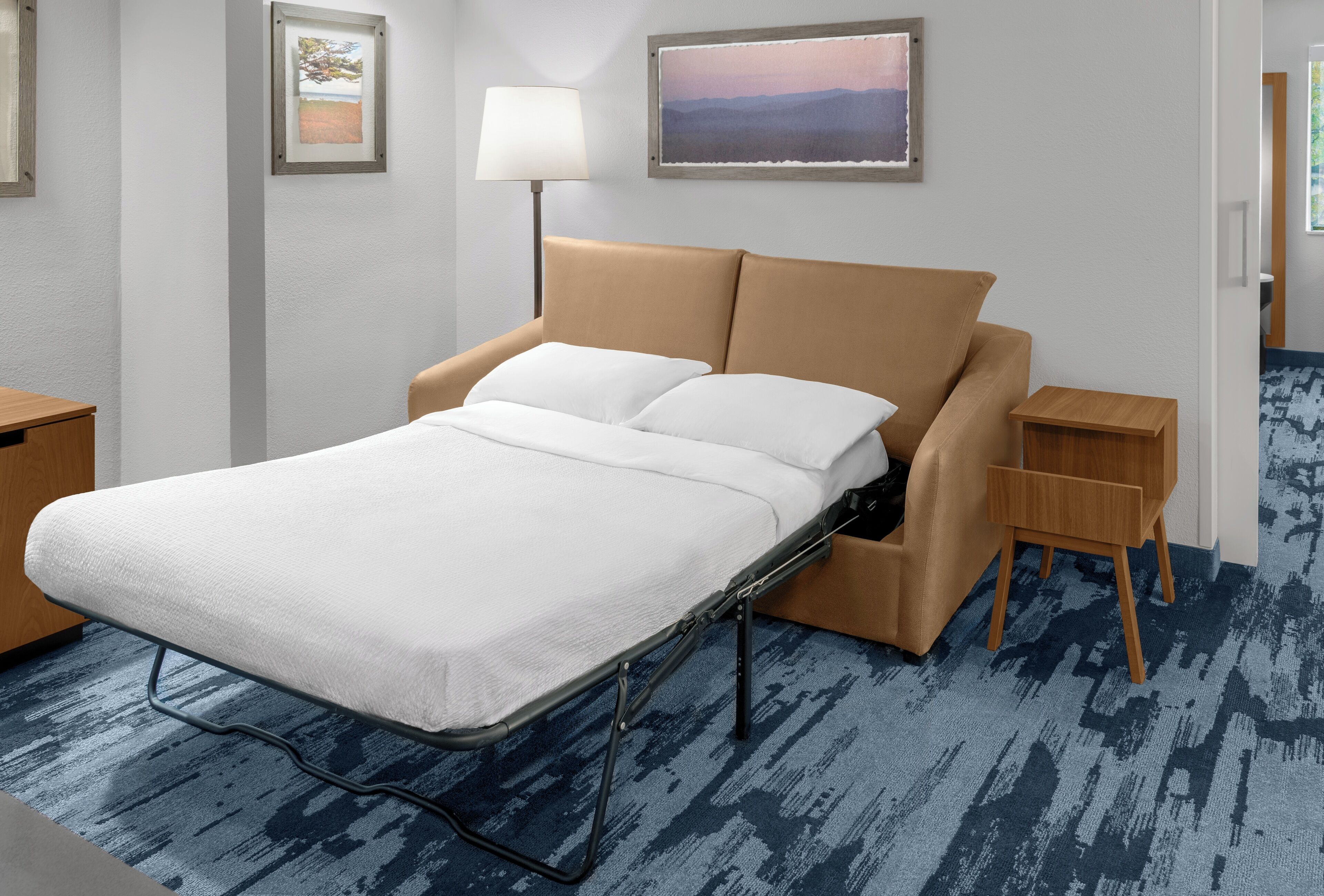 Une chambre d’hôtel avec un lit, une lampe de chevet, une petite table d’appoint en bois et un tableau encadré au mur.