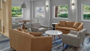Une chambre d’hôtel moderne avec un canapé beige, un fauteuil blanc et une table basse ronde. De grandes fenêtres offrent une vue sur la verdure et le paysage urbain.
