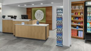 Un espace d’accueil d’hôtel comprenant un comptoir de réception en bois, un terminal d’enregistrement numérique, un point de distribution de boissons et une étagère à collations.