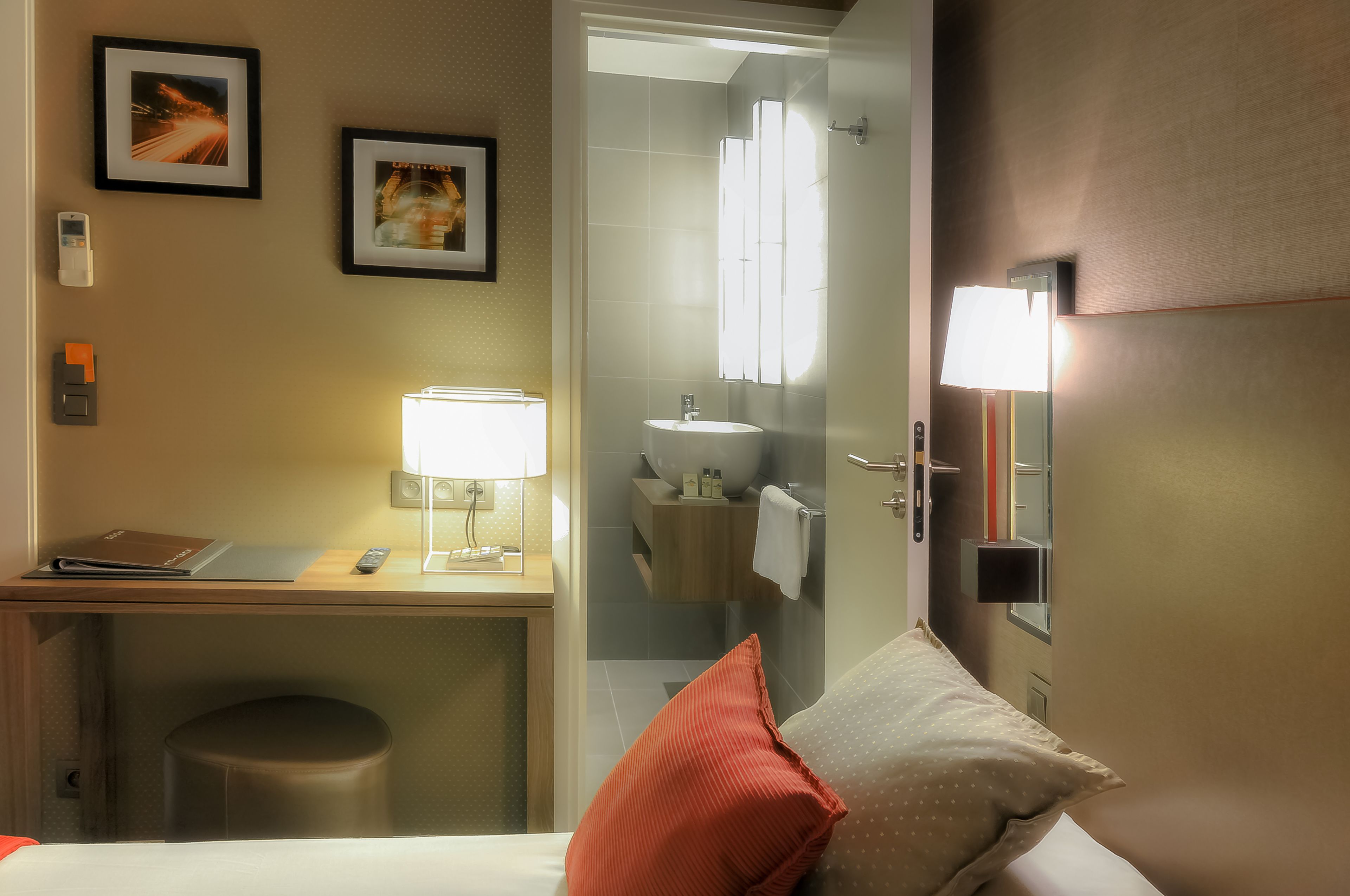 Hotelkamer met een bed, een bureau met lamp, een klein krukje, een badkamer met wastafel en spiegel, en lijstjes aan de muur.