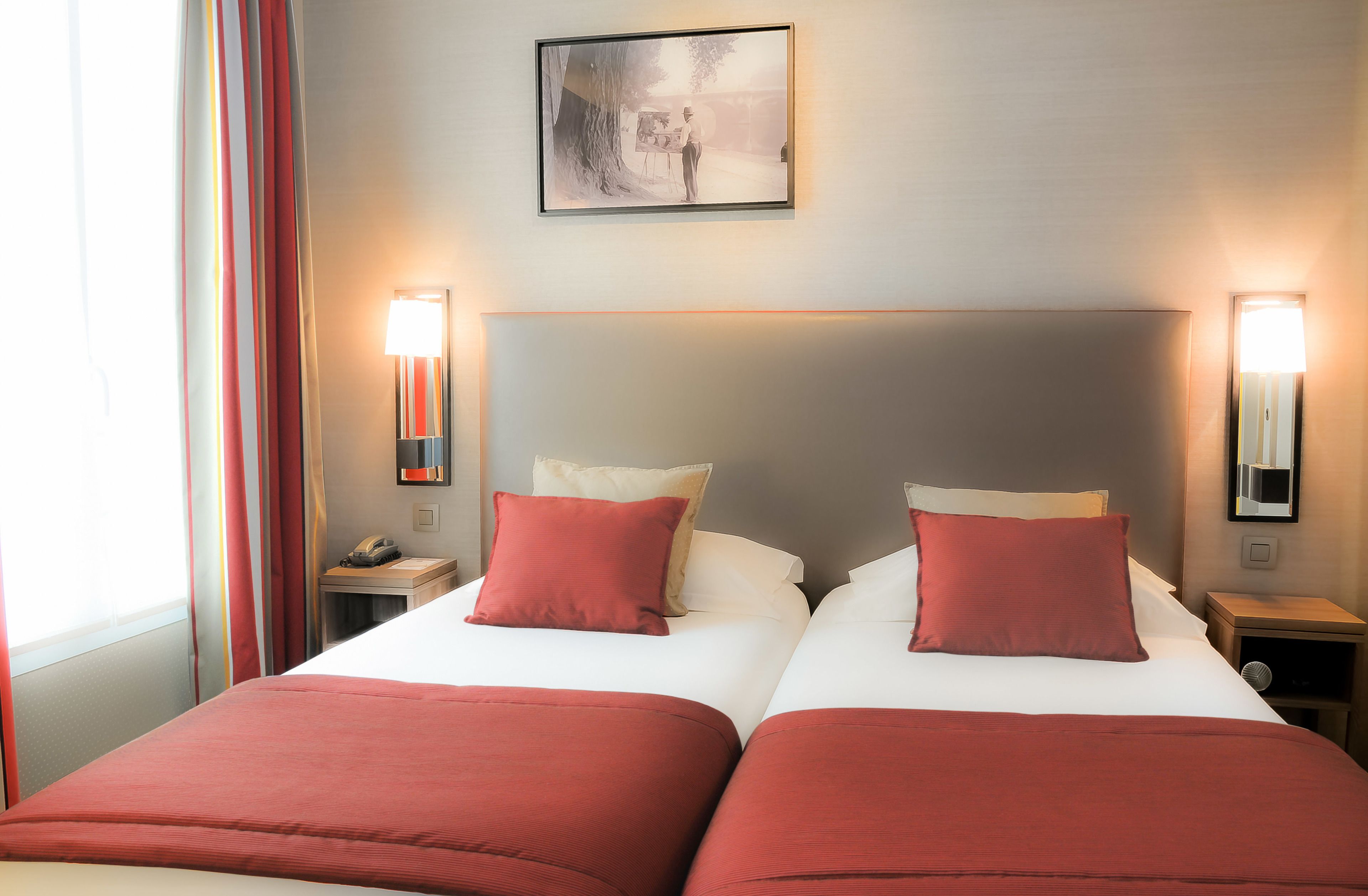 A double bed with red bedding, two bedside tables, a nightstand, a wall-mounted lamp, and a framed picture above the bed.