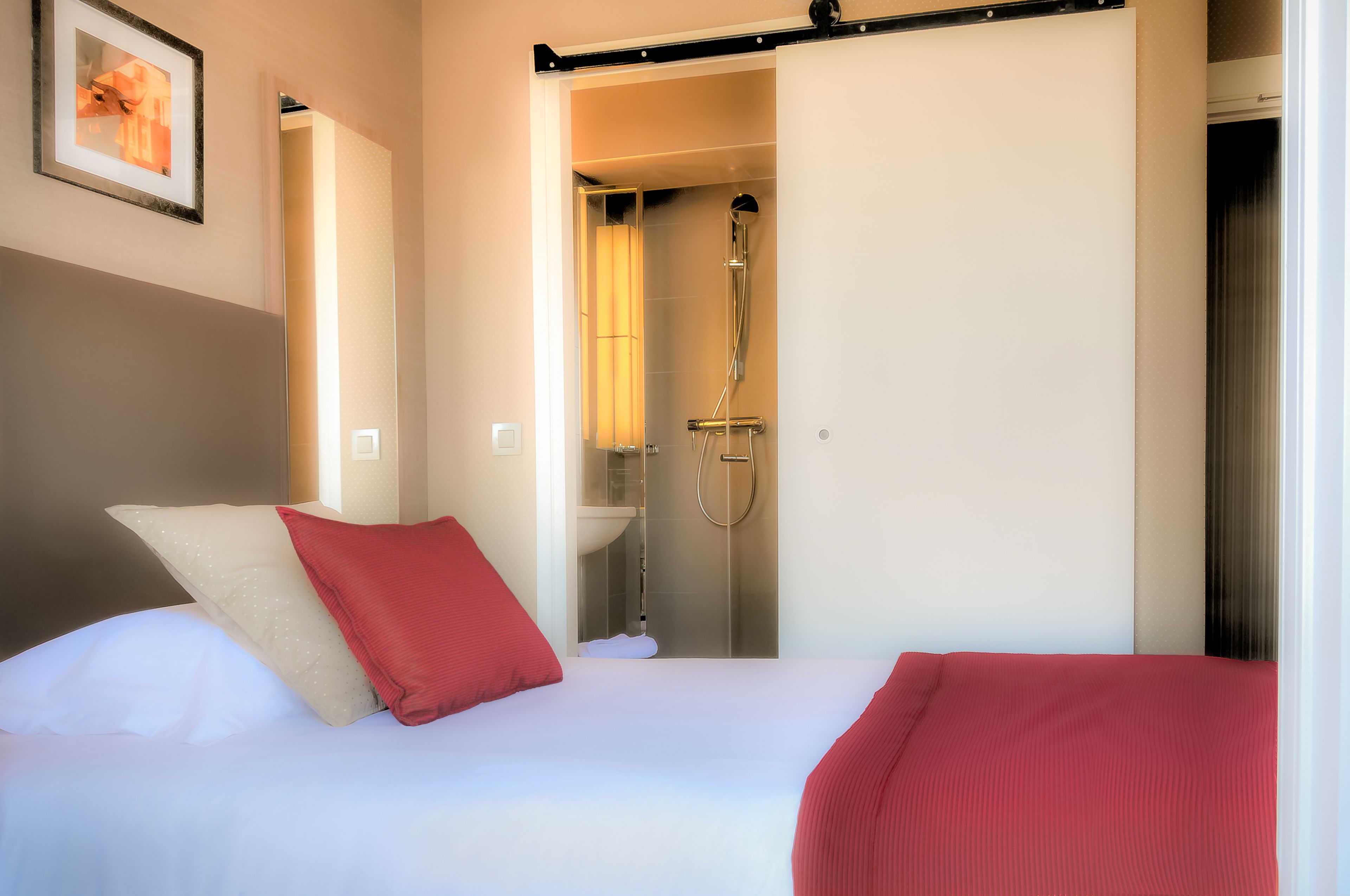 A hotel room with a bed, a red pillow, a white pillow, and a red blanket. There is a bathroom visible with a shower and a sink.