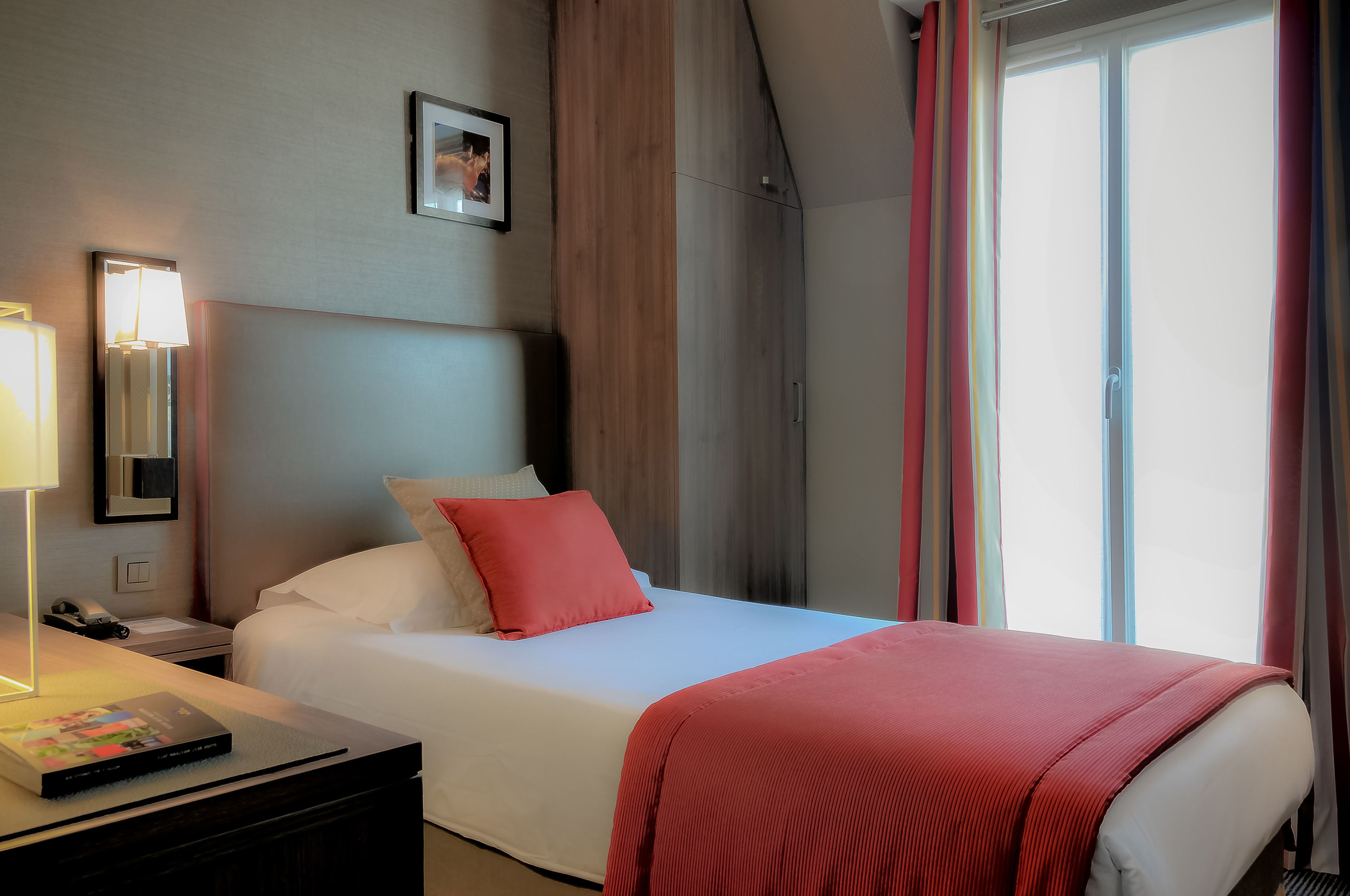 A hotel room with a bed, a bedside table with a lamp, a nightstand, and a window with curtains.