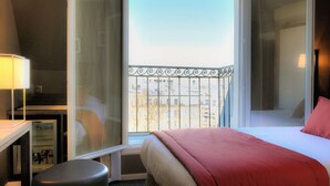 Quarto de hotel com cama, escrivaninha, cadeira, pequeno frigorífico e varanda com vista para edifícios.