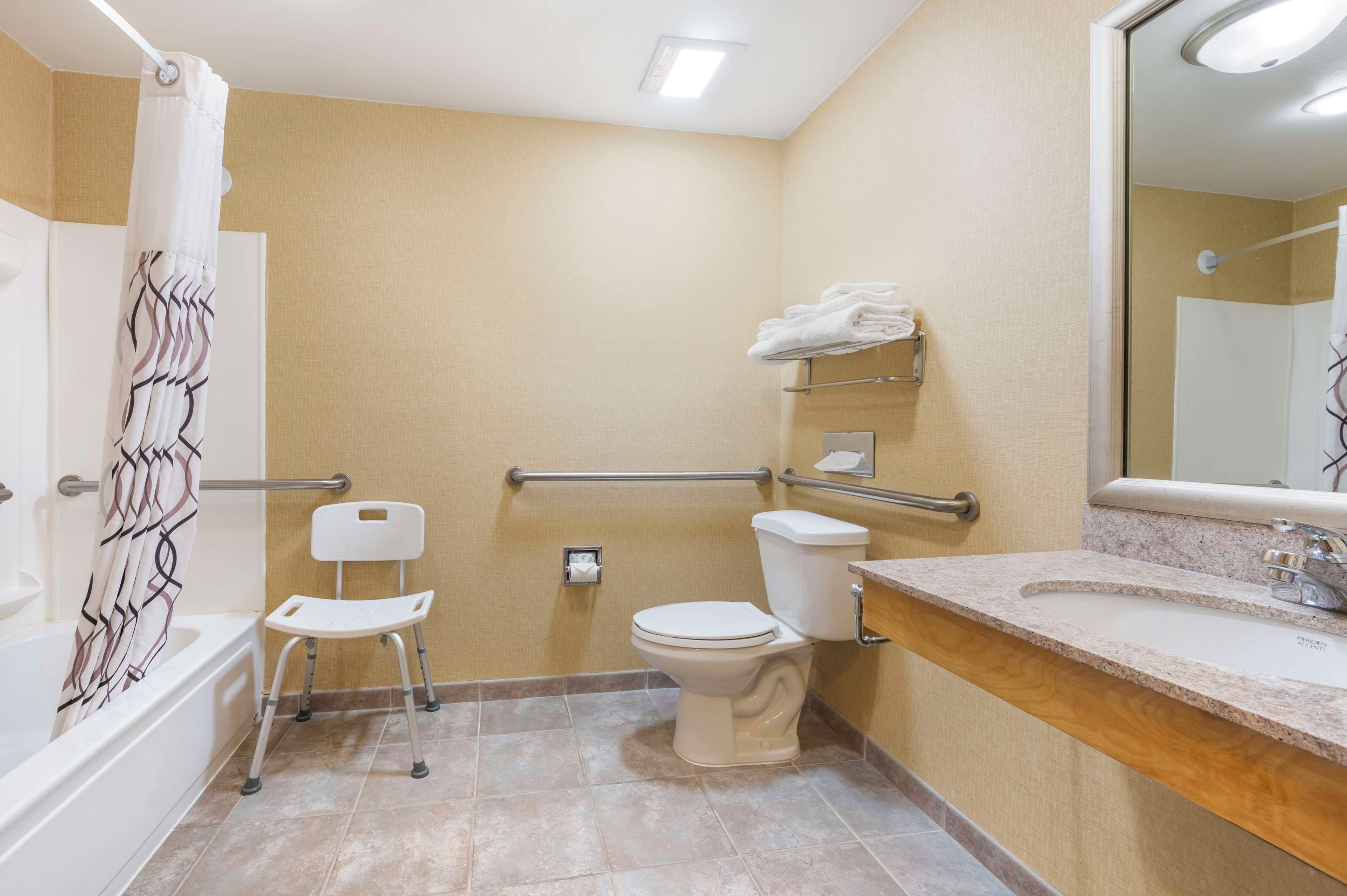 A bathroom with a shower, toilet, and sink. There is a chair and a handrail.