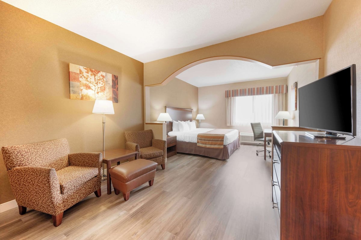 A hotel room with a bed, two armchairs, a desk with a TV, and a painting on the wall.
