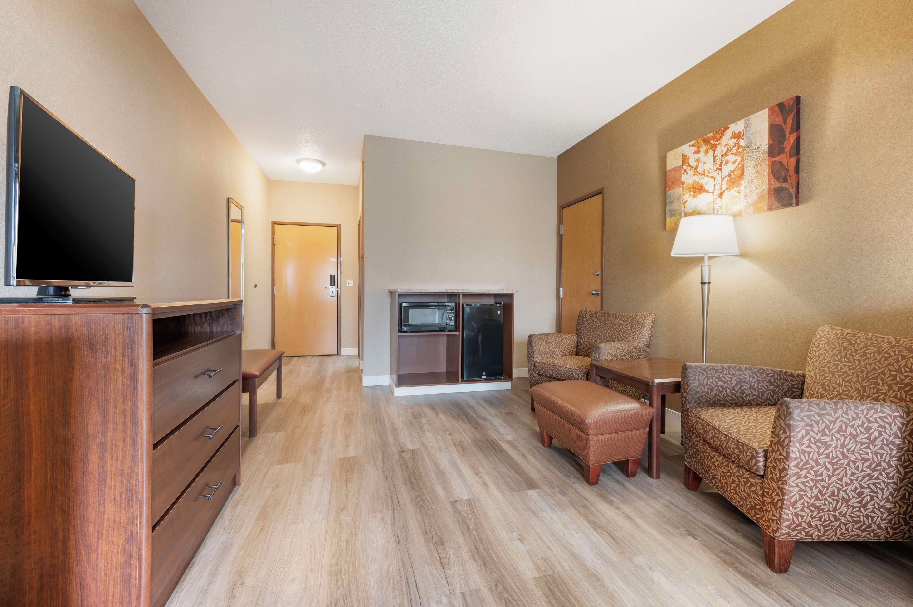 A hotel room with a flat-screen TV, a wooden dresser, a sofa, a chair, a table, a lamp, and a microwave.