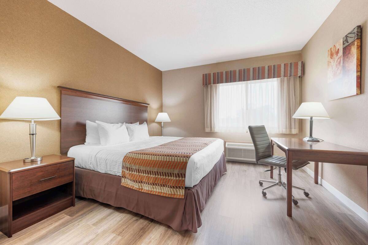 A hotel room with a large bed, two bedside tables with lamps, a desk with a chair, and a window with curtains.