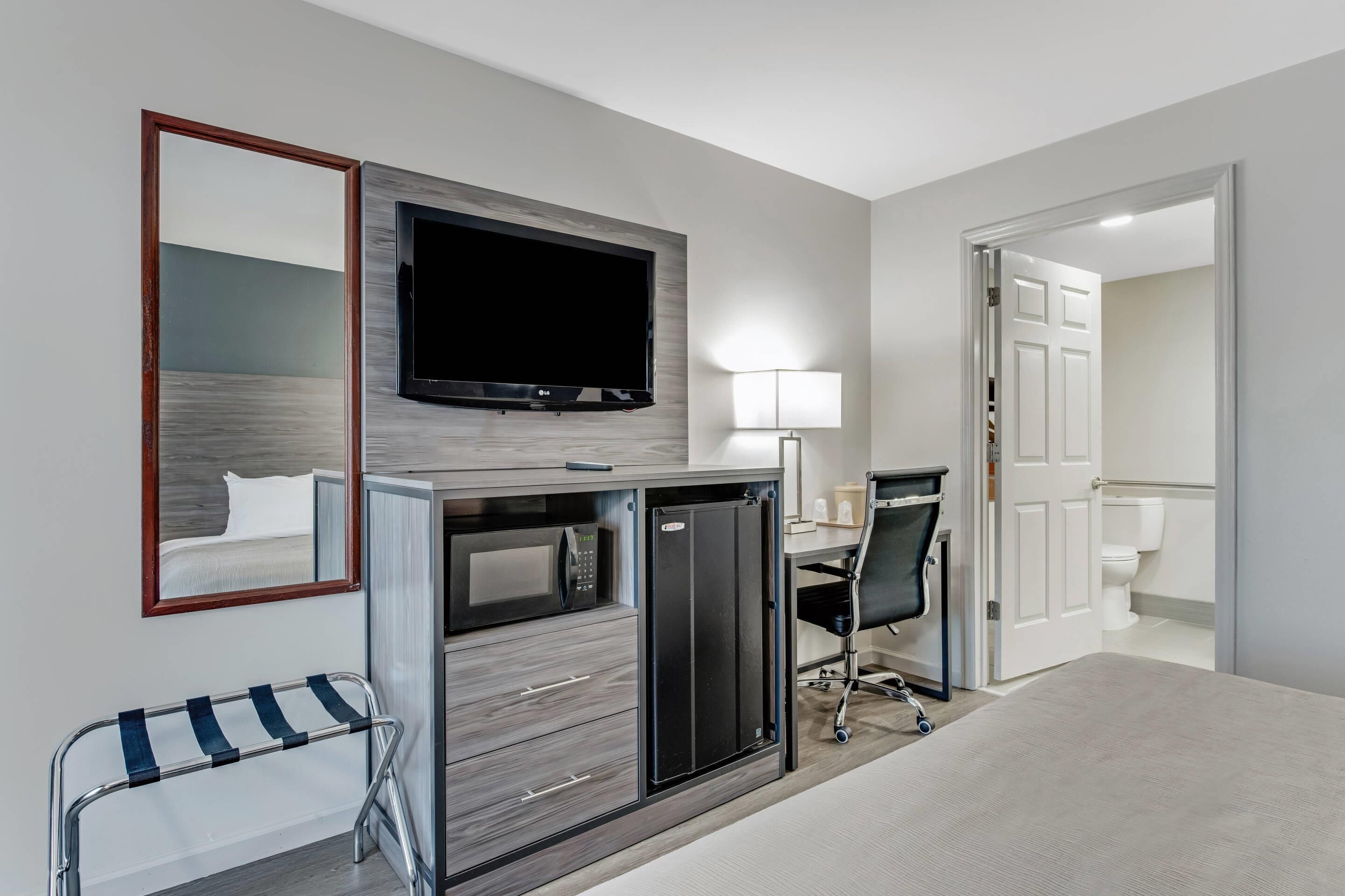 A hotel room with a flat-screen TV mounted on the wall, a desk with a chair, a microwave, and a mini-fridge.
