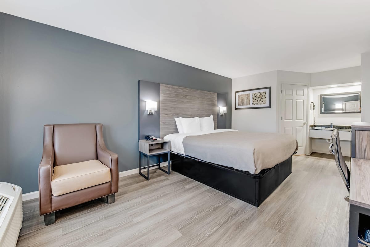 Une chambre d’hôtel moderne équipée d’un lit, d’une chaise, d’une table de chevet, d’un bureau et d’une salle de bain avec un miroir et un lavabo.