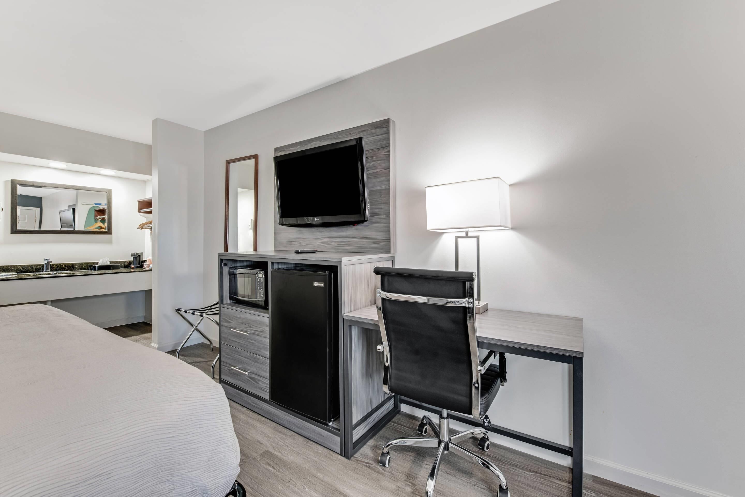 A modern hotel room with a bed, a desk with a chair, a flat-screen TV, a refrigerator, a microwave, a lamp, and a large mirror.