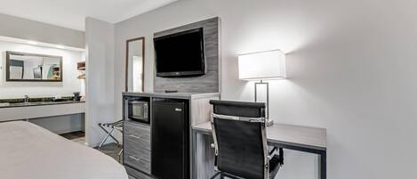 A modern hotel room with a bed, a desk with a chair, a flat-screen TV, a refrigerator, a microwave, a lamp, and a large mirror.