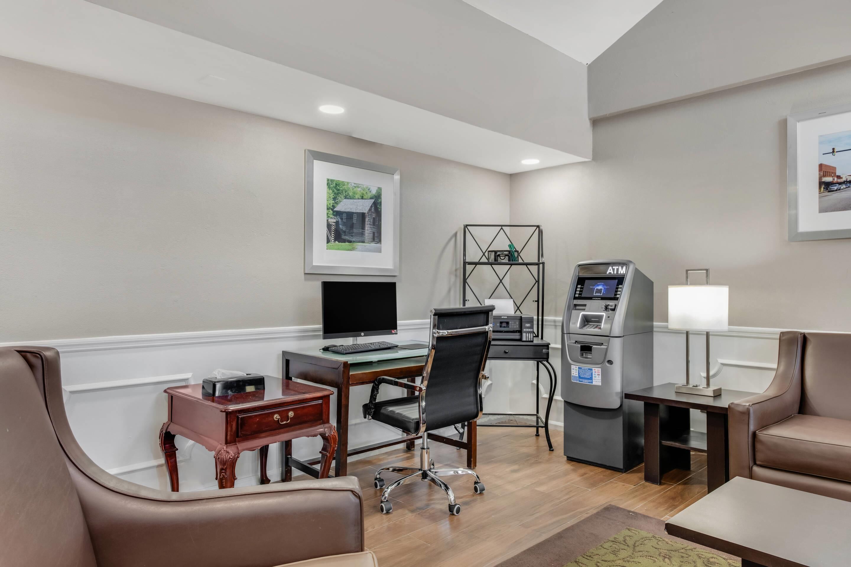 A hotel room with a desk, computer, chair, ATM, and a couch.
