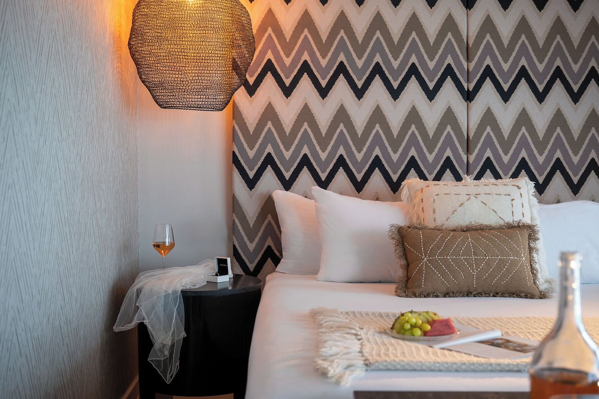A neatly arranged hotel room with a bed, a bedside table with a glass of wine, a decorative wall, and a textured wall on the left.