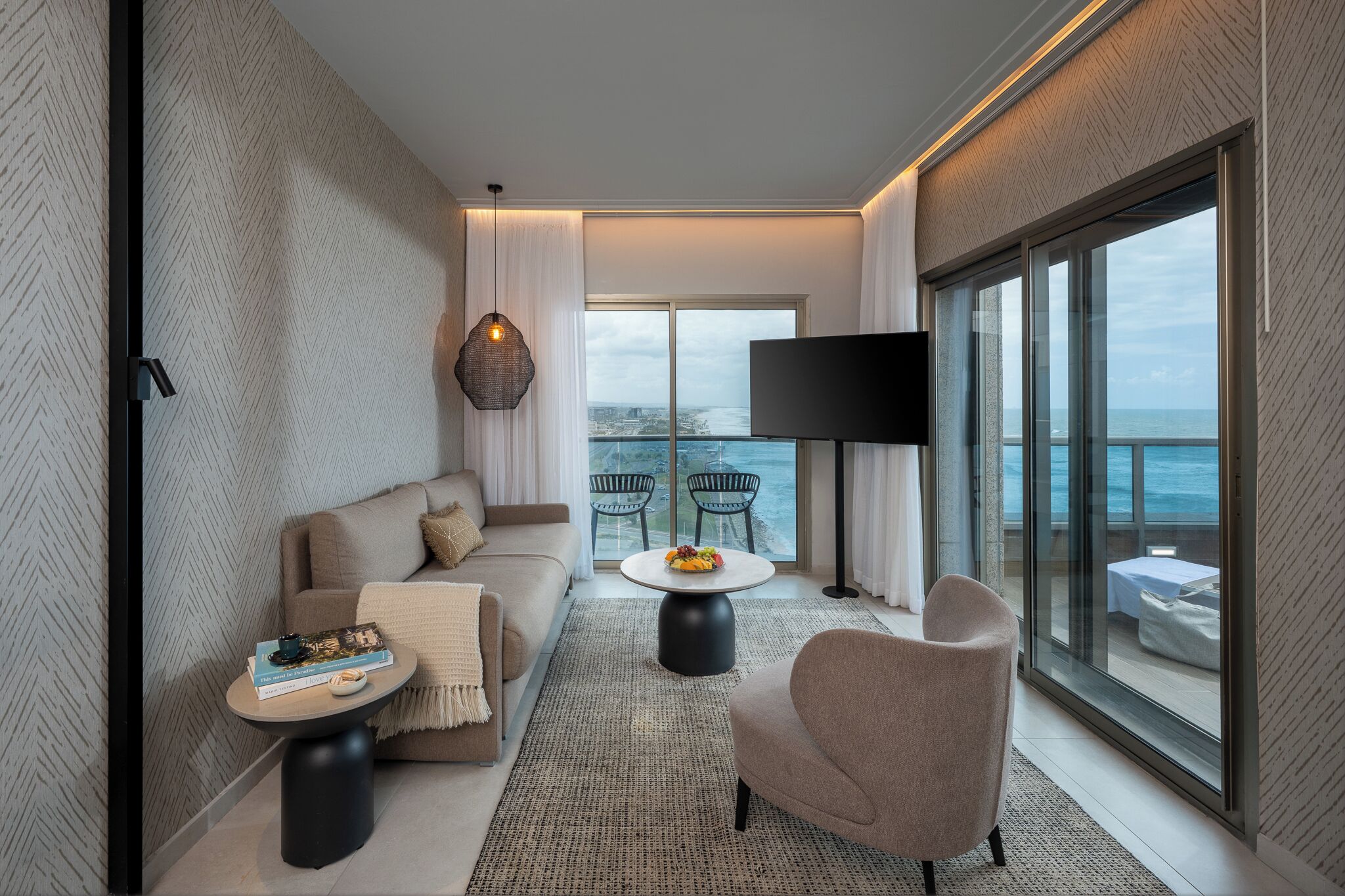 Een moderne woonkamer met een bank, fauteuil en salontafel, een flatscreen-tv en een balkon met uitzicht op zee.