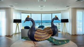 A room with a large peacock sculpture, a TV on a tripod stand, and a view of the sea through large windows.