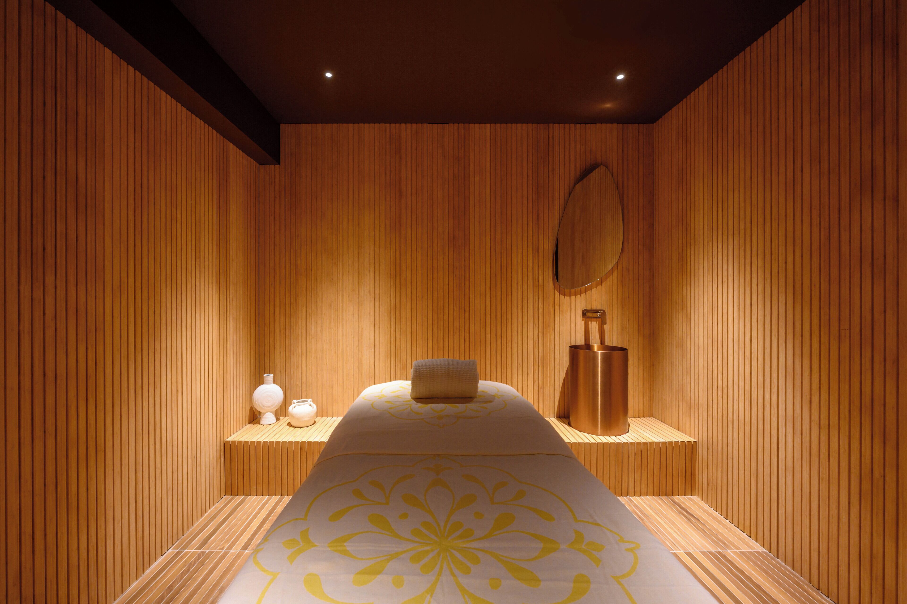A room with a wooden panel wall, a bed with a patterned bedspread, a bedside table with a vase, and a mirror.