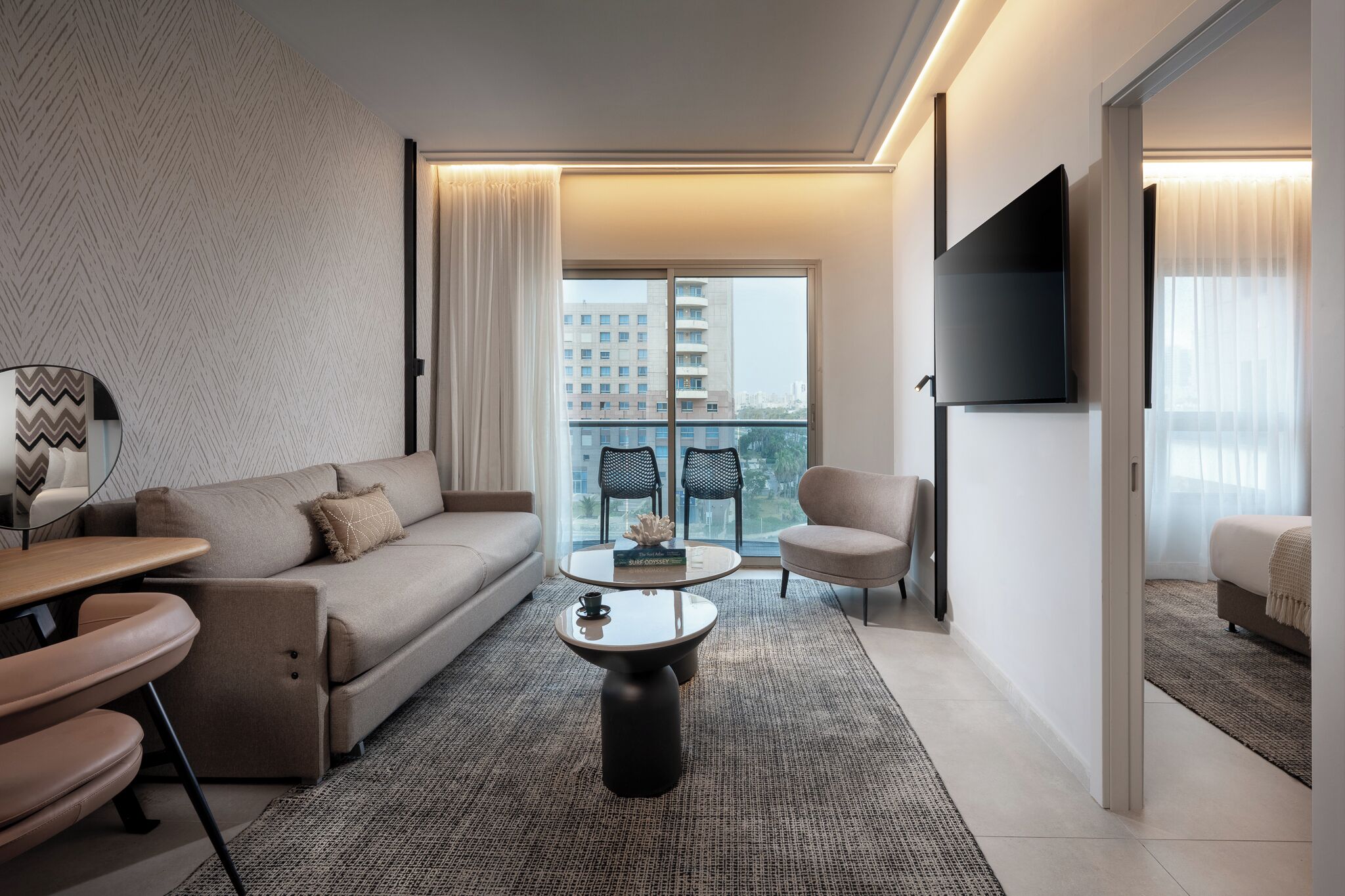 A modern hotel room with a sofa, a round coffee table, a TV mounted on the wall, and a view of the city through large windows.