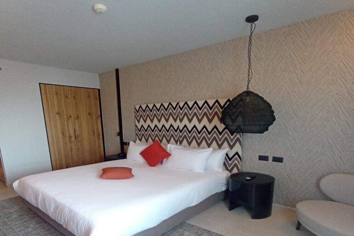 A modern hotel room with a large bed, a headboard with a zigzag pattern, a small black trash bin, and a grey sofa.