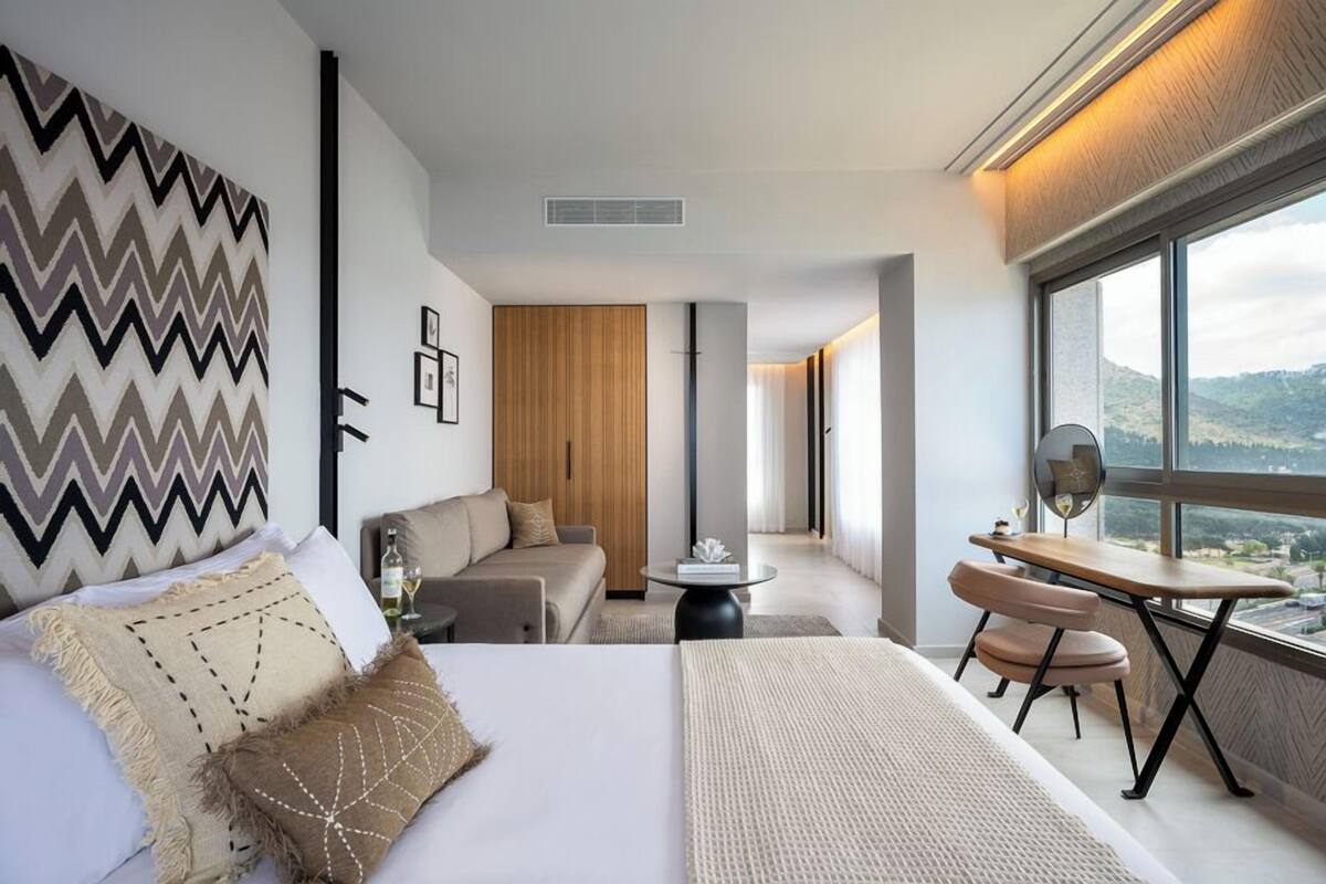 A modern hotel room with a large bed, a sofa, a dining table, and a view of the outdoors.