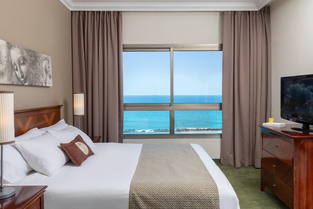 A hotel room with a large bed, a television, a desk, and a window with a view of the ocean.