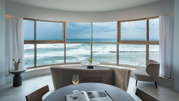 A room with a curved view of the ocean, a round table with a glass of wine, a chair, and a small table with a bottle and glasses. - Leonardo Plaza Haifa (Haifa)
