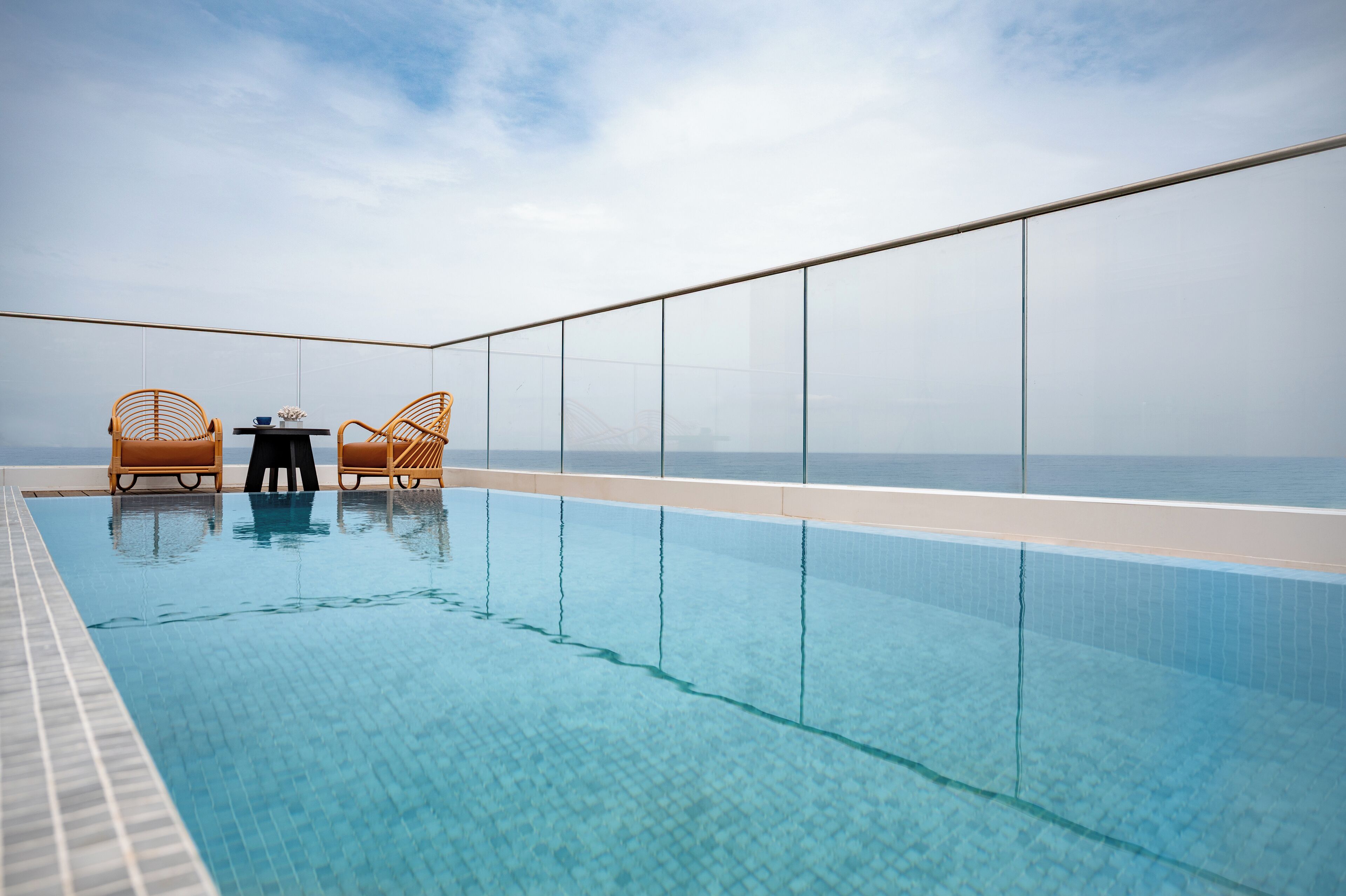 A rooftop infinity pool with clear blue water, two wicker chairs, and a table.