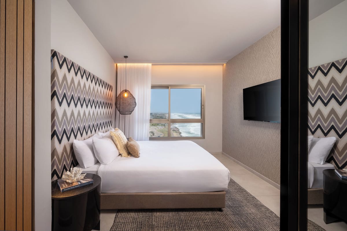 A modern hotel room with a large bed, a television mounted on the wall, and a view of the ocean through a large window.