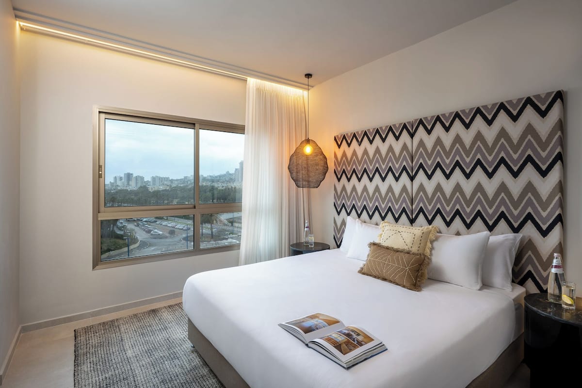 A hotel room with a large bed, a view of the city, a patterned headboard, and a magazine on the bed.