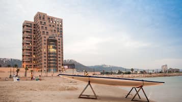 Una playa con un edificio grande al fondo, una orilla arenosa y un bote sobre un soporte.