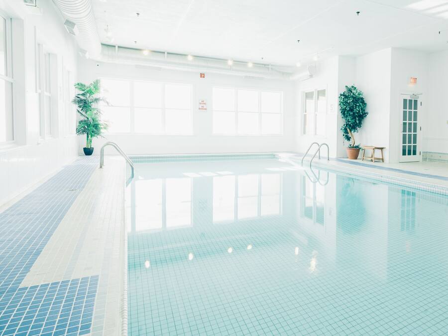 An indoor swimming pool with clear blue water, tiled floor, and potted plants.