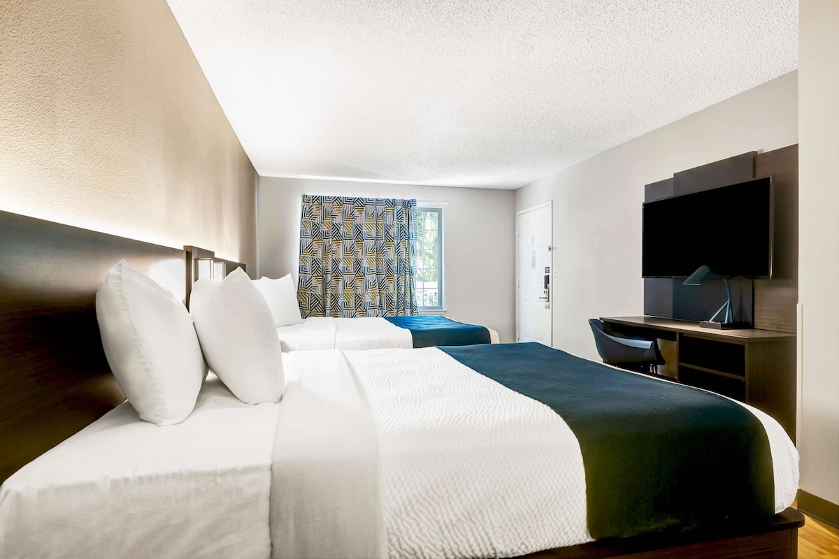 A hotel room with a large bed, a desk with a chair, a flat-screen TV, and a window with patterned curtains.