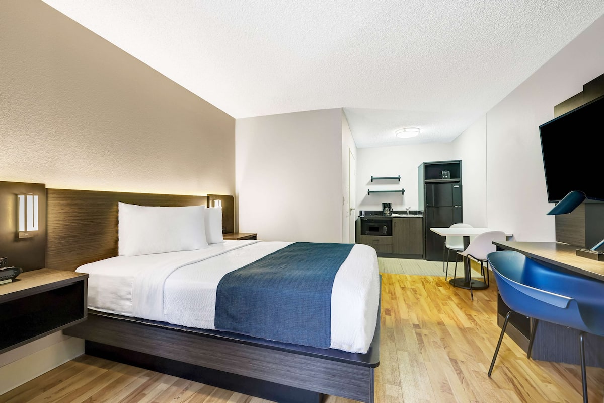 A modern hotel room with a bed, desk, and kitchenette.