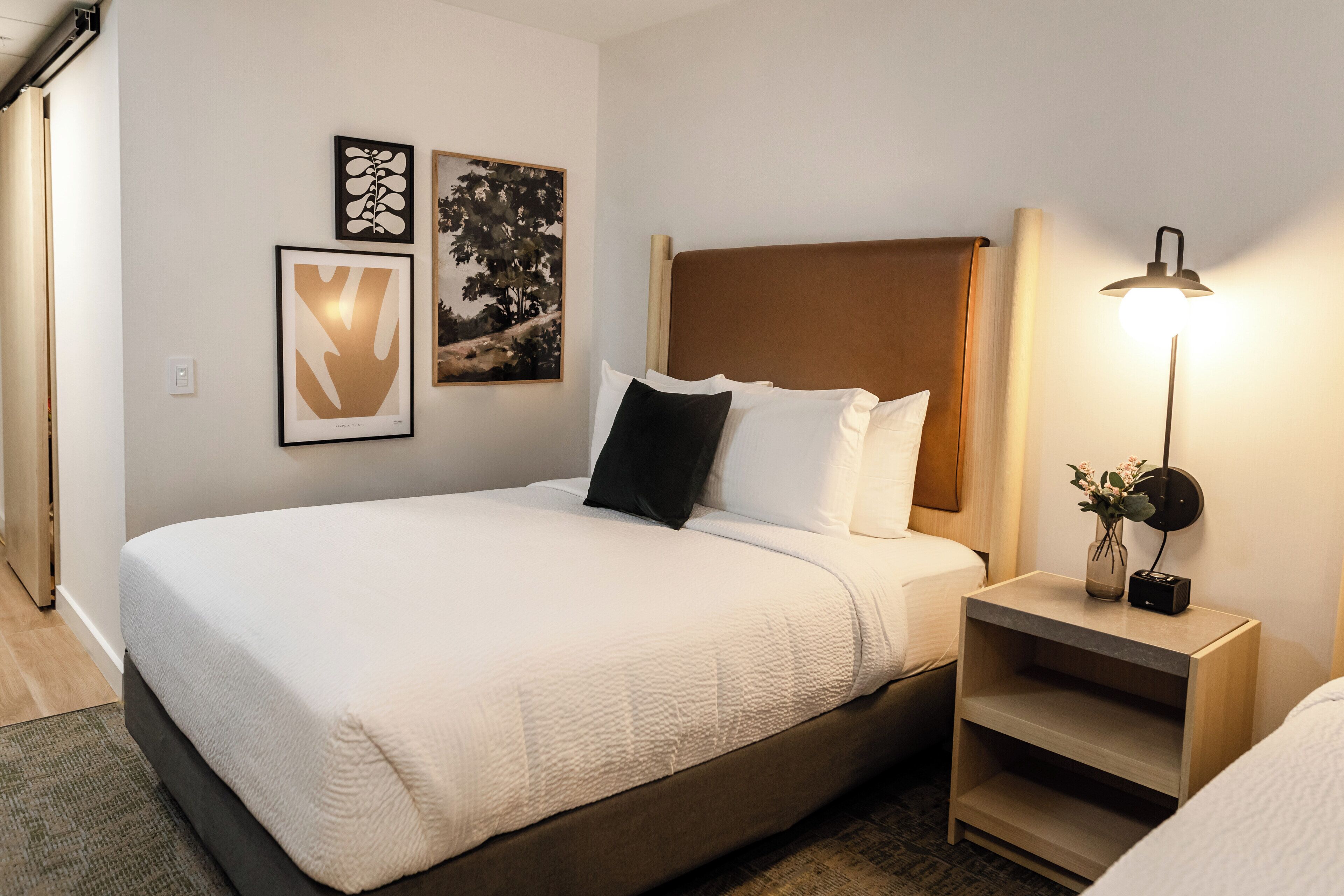 A neatly made bed with a white comforter and pillows, a bedside table with a lamp, and two framed artworks on the wall.