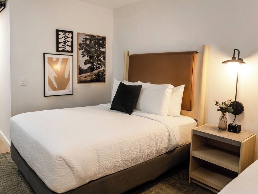 A neatly made bed with a white comforter and pillows, a bedside table with a lamp, and two framed artworks on the wall.