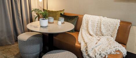 A cozy living area with a round table, two ottomans, a brown leather sofa with a white throw blanket, and a potted plant.