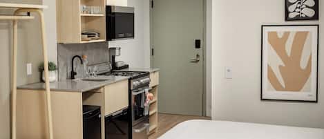 A compact kitchen with a stove, sink, and cabinets, a bed with a white bedspread, and two framed artworks on the wall.