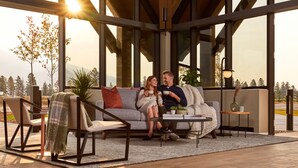 A modern living room with a sofa, chairs, and a coffee table. There are large windows with a view of trees and mountains.