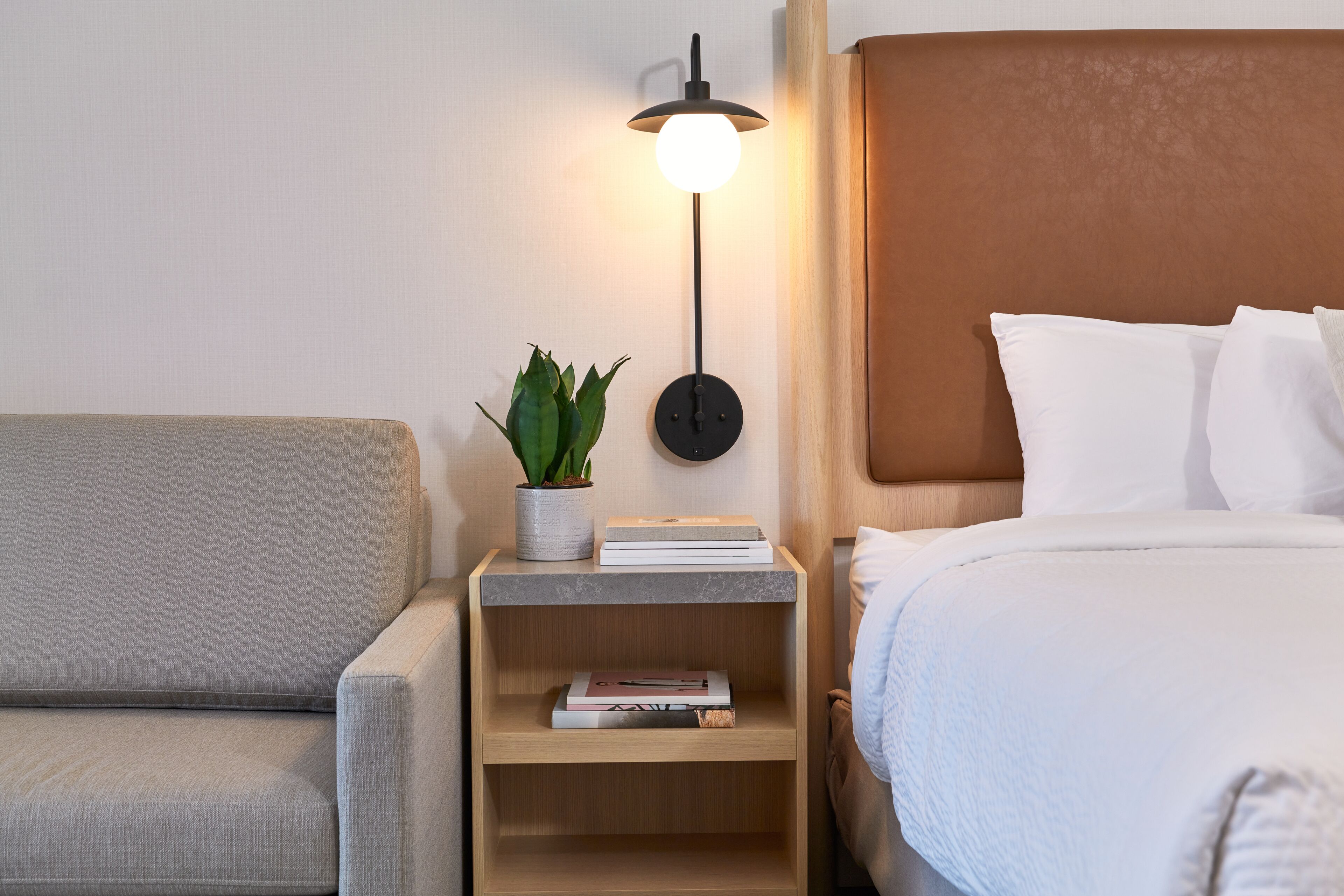 A neatly arranged hotel room with a bed, a bedside table with a lamp, a small plant, and a few books.